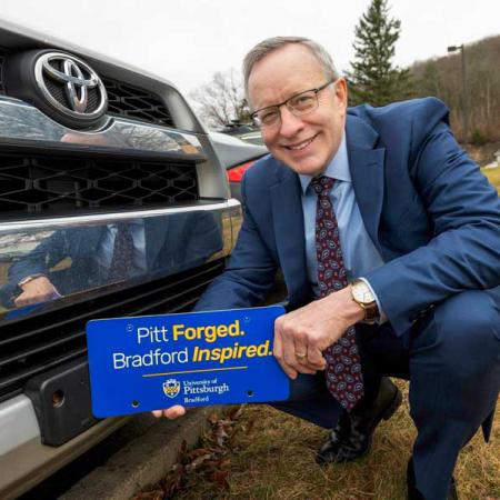 President Rick Esch in front of a vehicle holding a license plate that says "Pitt Forged. Bradford Inspired"
